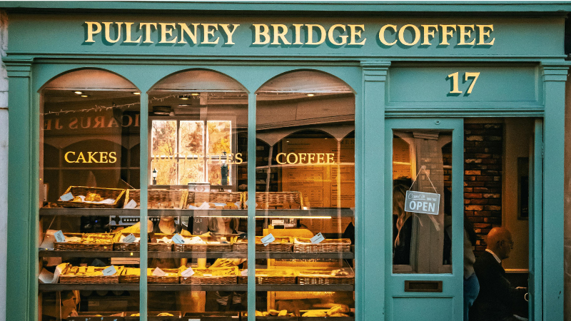 Kaféet Pulteney Bridge Coffee i Bath, England, med grön fasad och skyltfönster fyllt med bakverk – populärt stopp vid Pulteney Bridge.